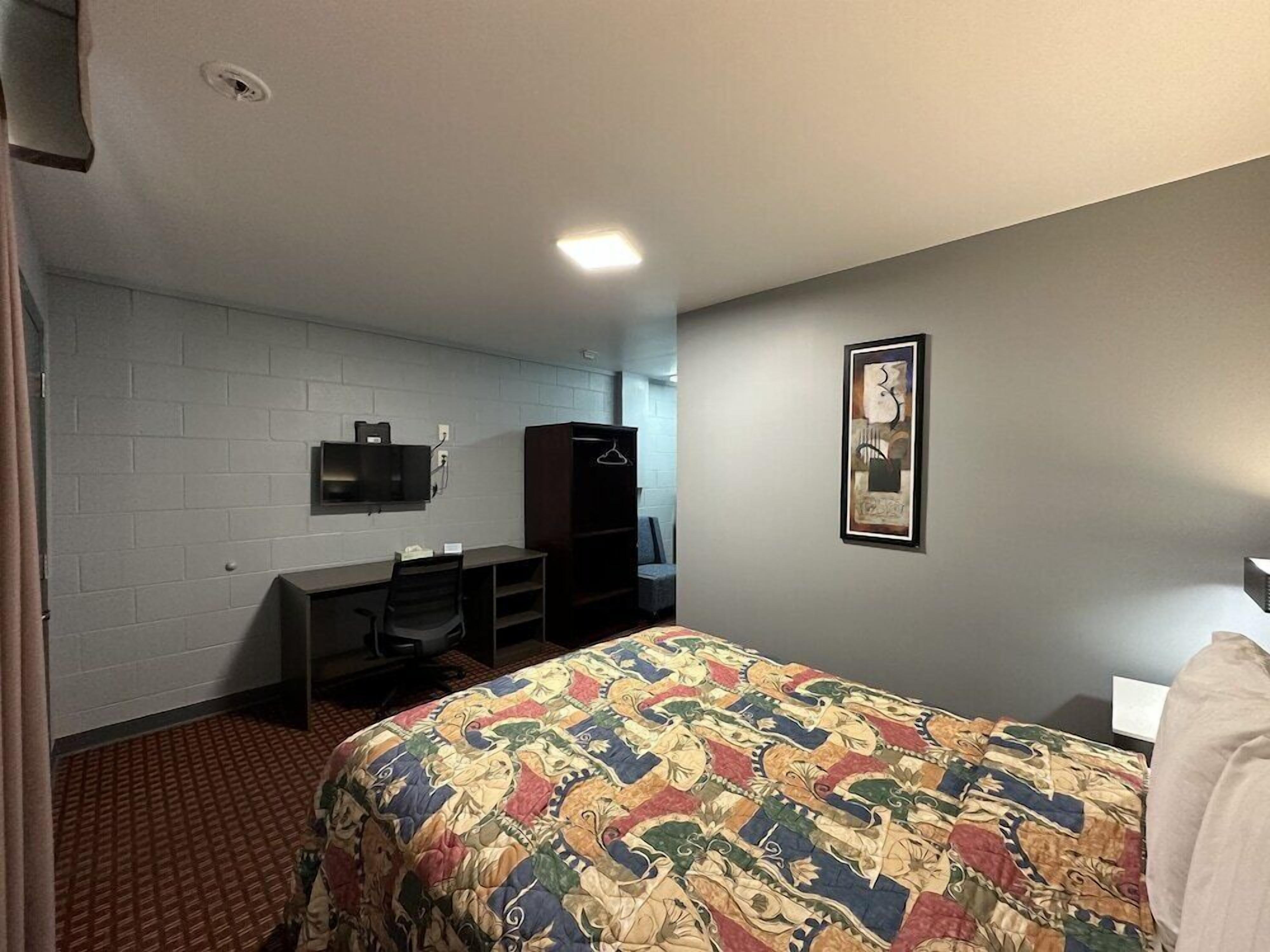 A hotel room with a bed, a desk, a chair, a wardrobe, a television mounted on the wall, and a framed artwork on the wall.