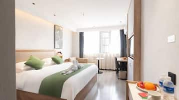 Habitación de hotel con cama, escritorio con silla, una pequeña mesa con fruta y una botella de agua, y ventana con cortinas.