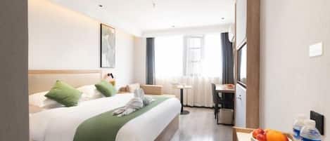A hotel room with a bed, a desk with a chair, a small table with fruit and a water bottle, and a window with curtains.