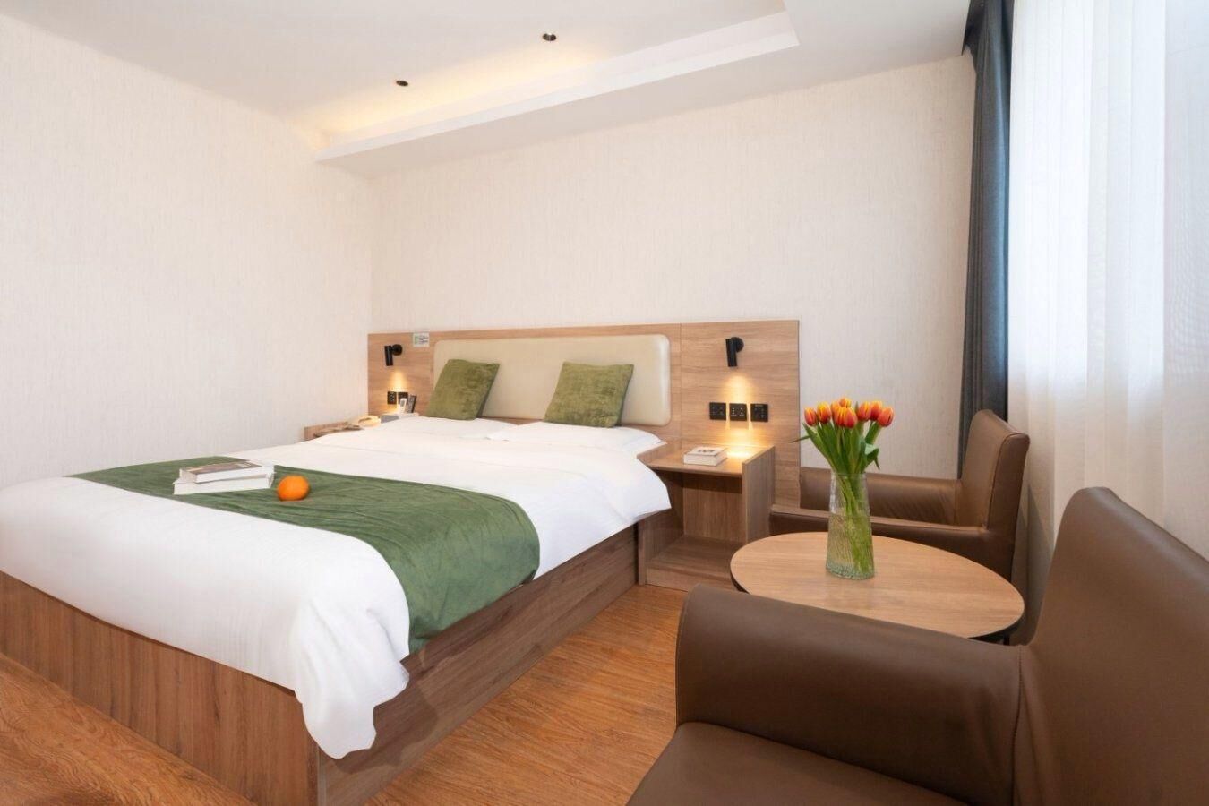 A hotel room with a large bed, a wooden headboard, a bedside table with a lamp, a small table with a vase of flowers, and a brown leather chair.