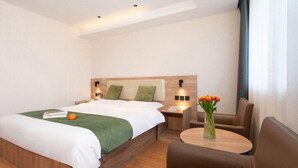 Una habitación de hotel con una cama grande, un cabecero de madera, una mesita de noche con una lámpara, una pequeña mesa con un florero y un sillón de cuero marrón.