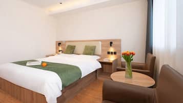A hotel room with a large bed, a wooden headboard, a bedside table with a lamp, a small table with a vase of flowers, and a brown leather chair.