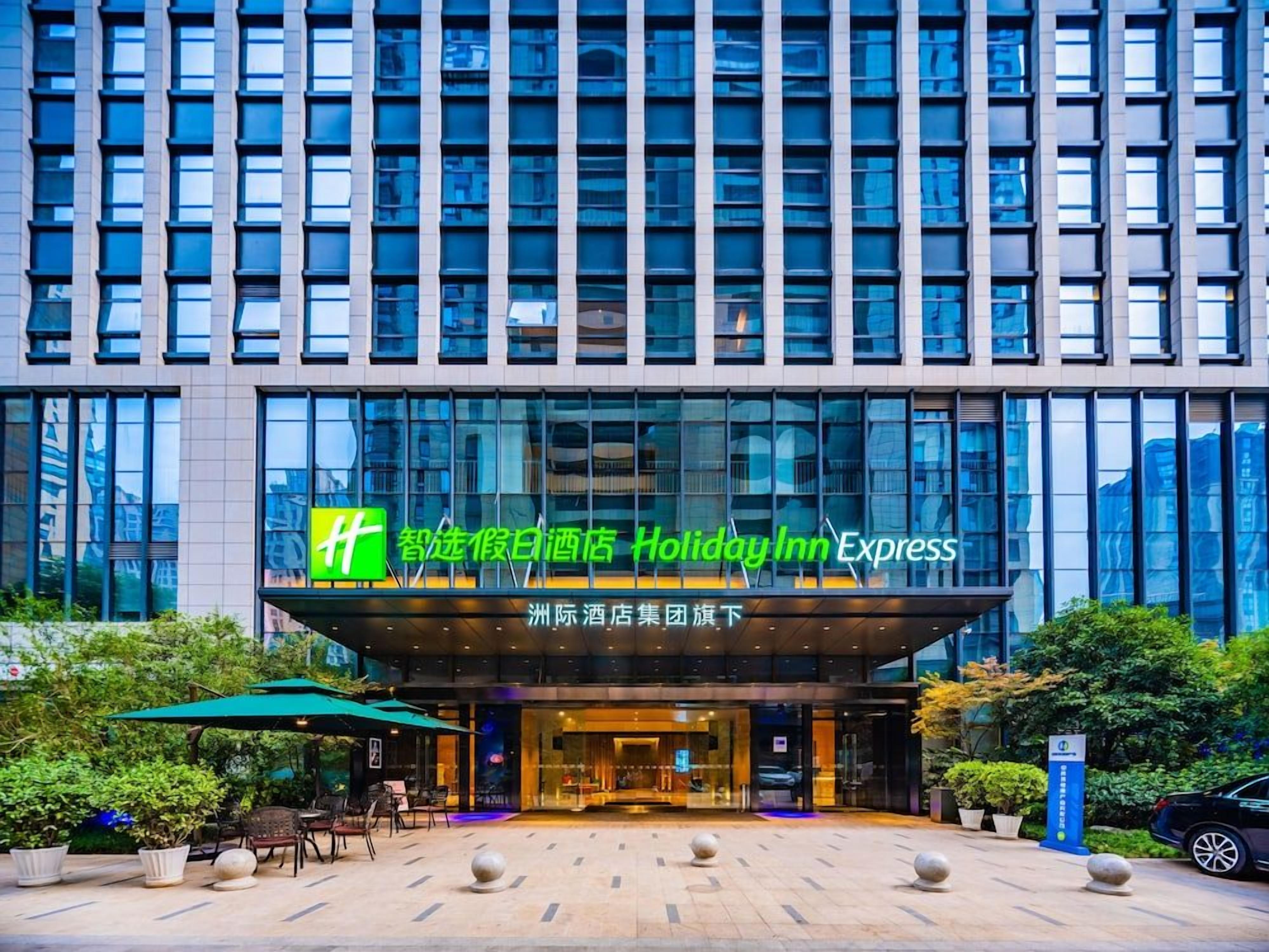 Un hôtel Holiday Inn Express doté d’une façade moderne, de grandes fenêtres et d’un espace extérieur aménagé pour s’asseoir.
