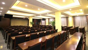 A conference room with rows of wooden tables and chairs, recessed ceiling lighting, and a stage area.