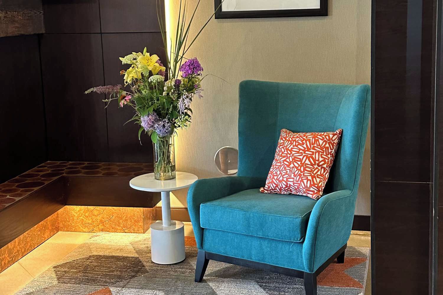 A teal armchair with a patterned pillow, a small round table with a vase of flowers, and a patterned rug.