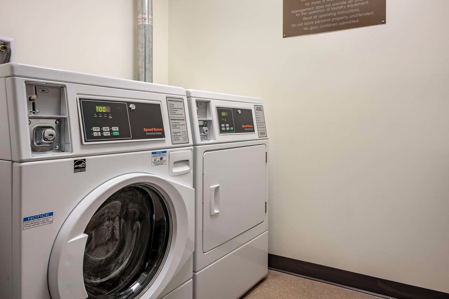 A row of white commercial washers with digital displays, a notice sign, and a safety sign on the wall.