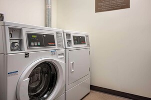 Una hilera de lavadoras industriales blancas con pantallas digitales, un cartel informativo y un letrero de seguridad en la pared.