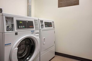 A row of white commercial washers with digital displays, a notice sign, and a safety sign on the wall. - Comfort Inn Connellsville Riverview (Connellsville)