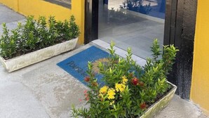 Un bâtiment jaune avec une grande porte vitrée, une terrasse en bois et des plantes en pot devant l’entrée.