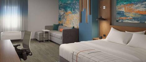 A modern hotel room with a large bed, a desk, a chair, and a sofa. There are two abstract paintings on the walls.