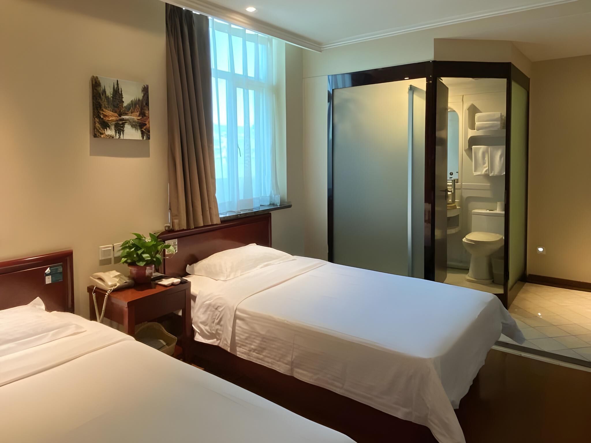 A hotel room with two beds, a window with curtains, a painting, a plant, a phone, and a glass-enclosed bathroom.
