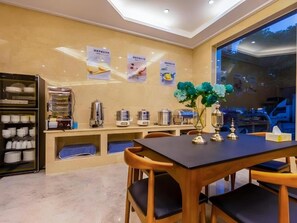 A hotel dining area with a table, chairs, a vase with flowers, and a counter with kitchen equipment. - Shell Hotel (Wuxi)