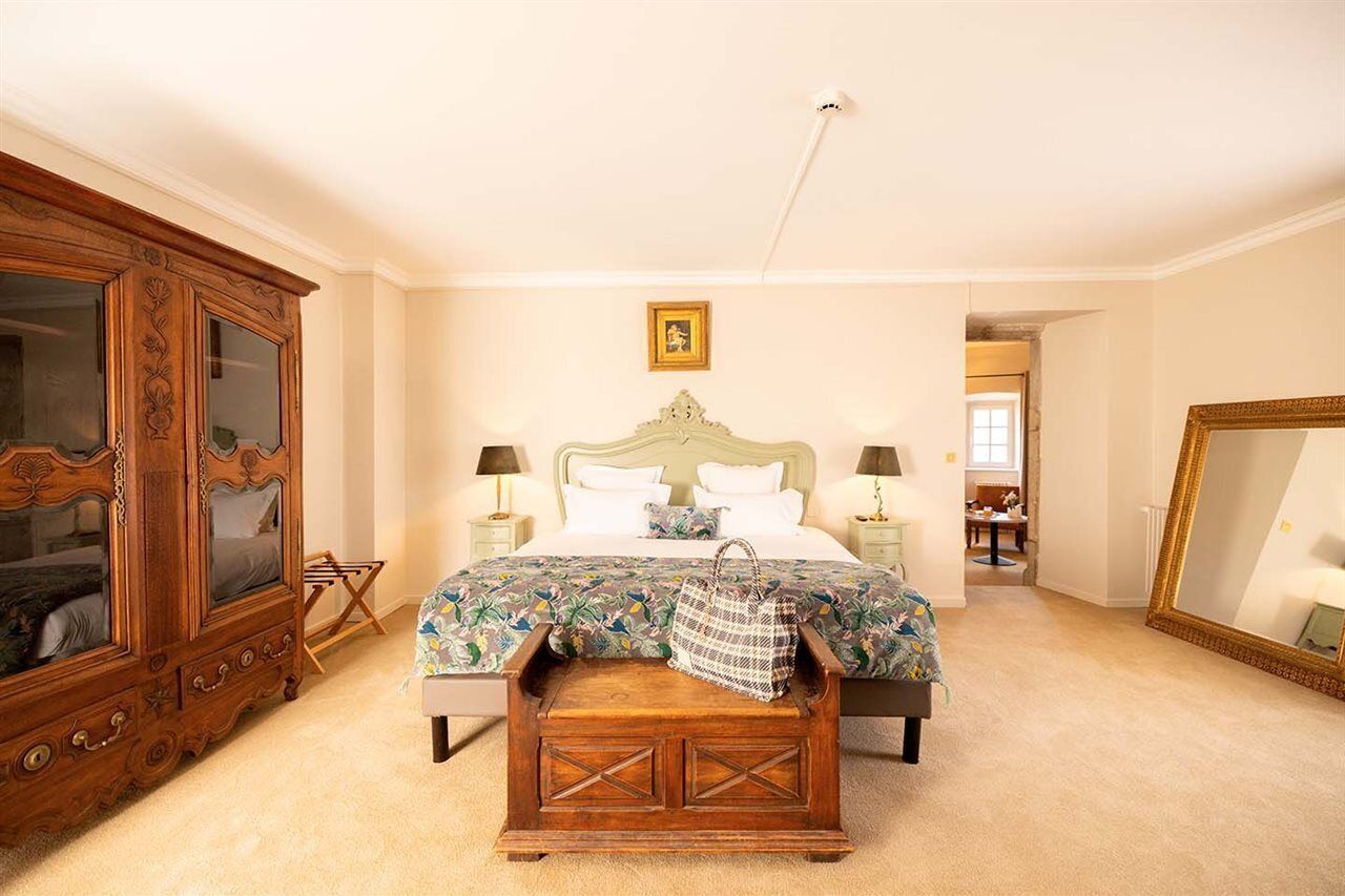 Une chambre avec une grande armoire en bois, un lit recouvert d’une couvre-lit à motifs floraux, une commode en bois et un grand miroir accroché au mur.