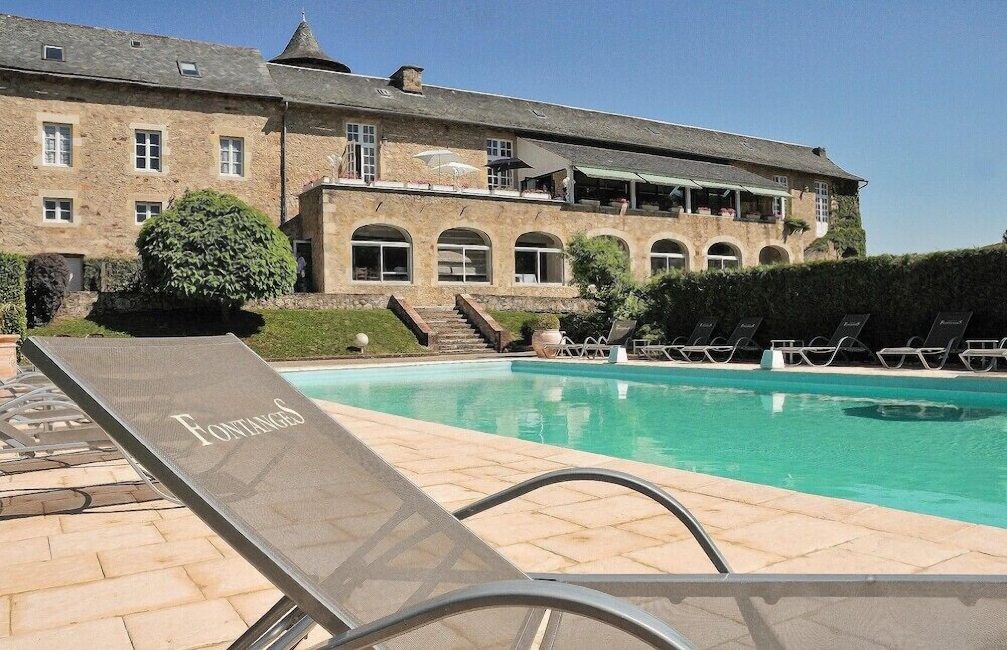 Un espace piscine avec des chaises longues et un bâtiment doté de fenêtres en arc de cercle et d’un balcon.