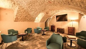 A cozy room with a stone ceiling, green upholstered chairs, a flat-screen TV, and a small table with a chessboard.