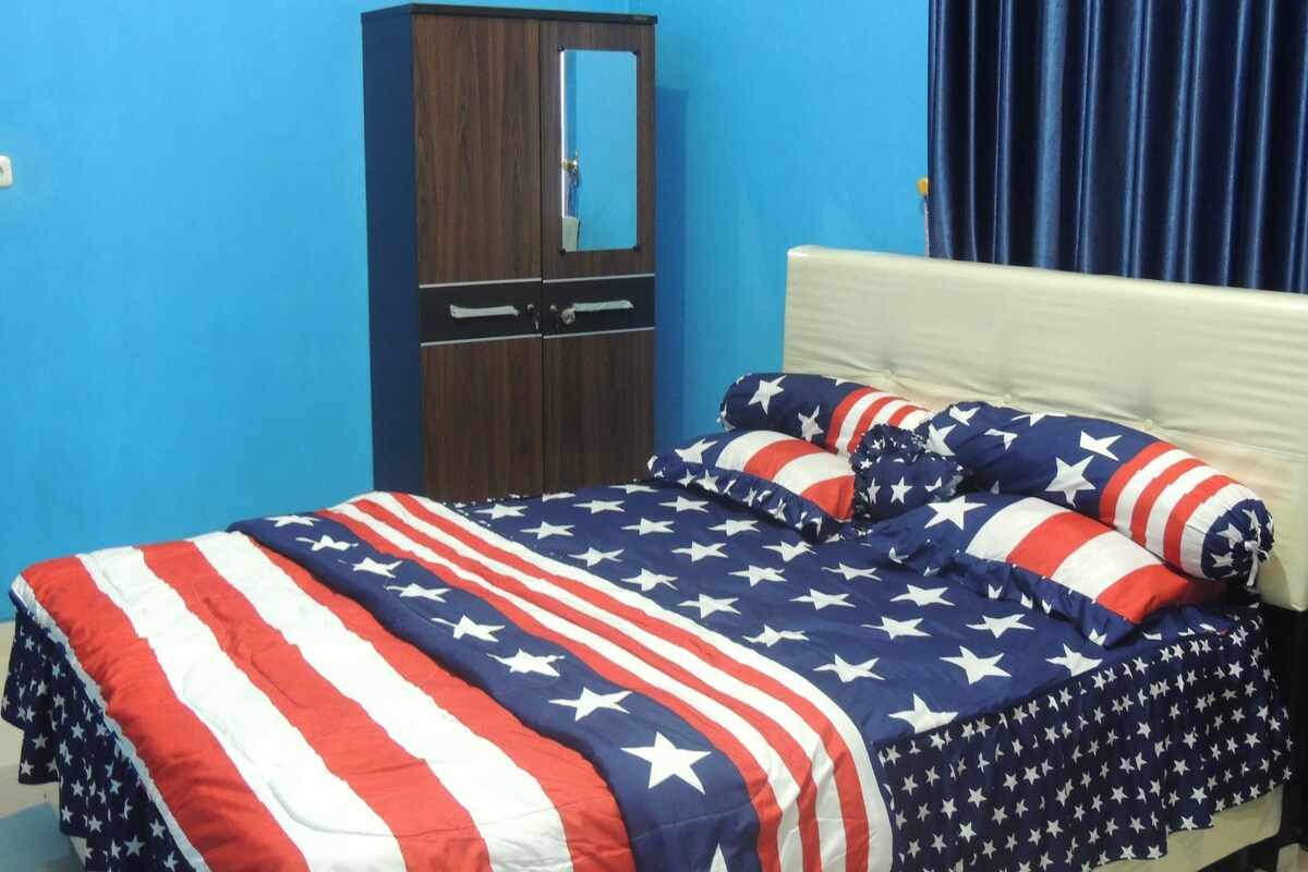 Un dormitorio con una cama cubierta con una colcha temática de la bandera estadounidense, un armario de madera y una cortina azul.