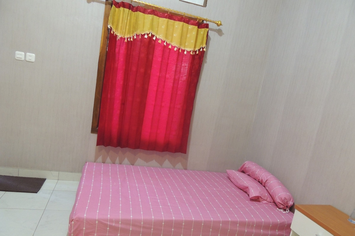 Un dormitorio con cama, una mesita de noche, una ventana con cortina e interruptor de luz en la pared.