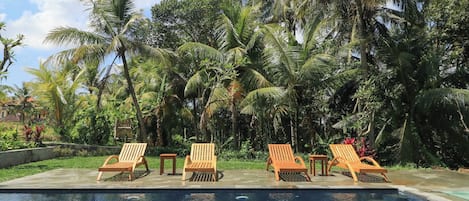 A poolside area with wooden lounge chairs and a clear blue pool, surrounded by lush palm trees.