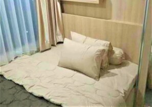 Una cama con edredón y almohadas color crema, un cabecero de madera y una ventana con cortinas.