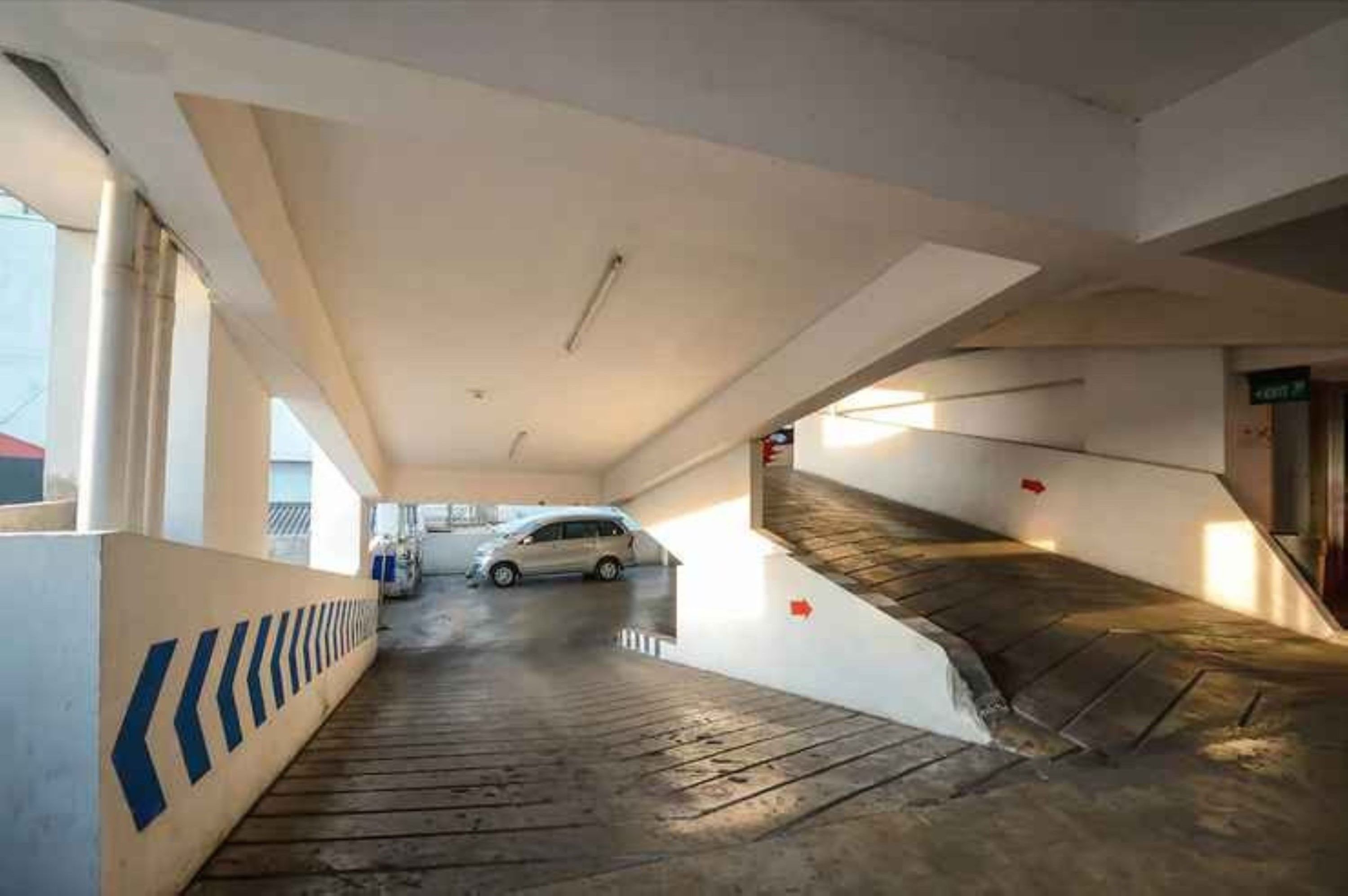 A parking garage with a white car parked in the distance, a ramp leading to an exit, and a blue and white striped safety line along the wall.