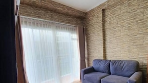 A living room with a sofa, wooden flooring, a stone wall, and a window with curtains. - Gateway Pasteur Apartment Bandung (Cimahi)
