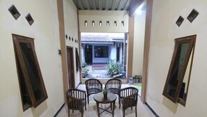 A corridor with a tiled floor, a round table with chairs, and a view of a building with a tiled roof and plants outside. - Homestay Sekar (Borobudur)
