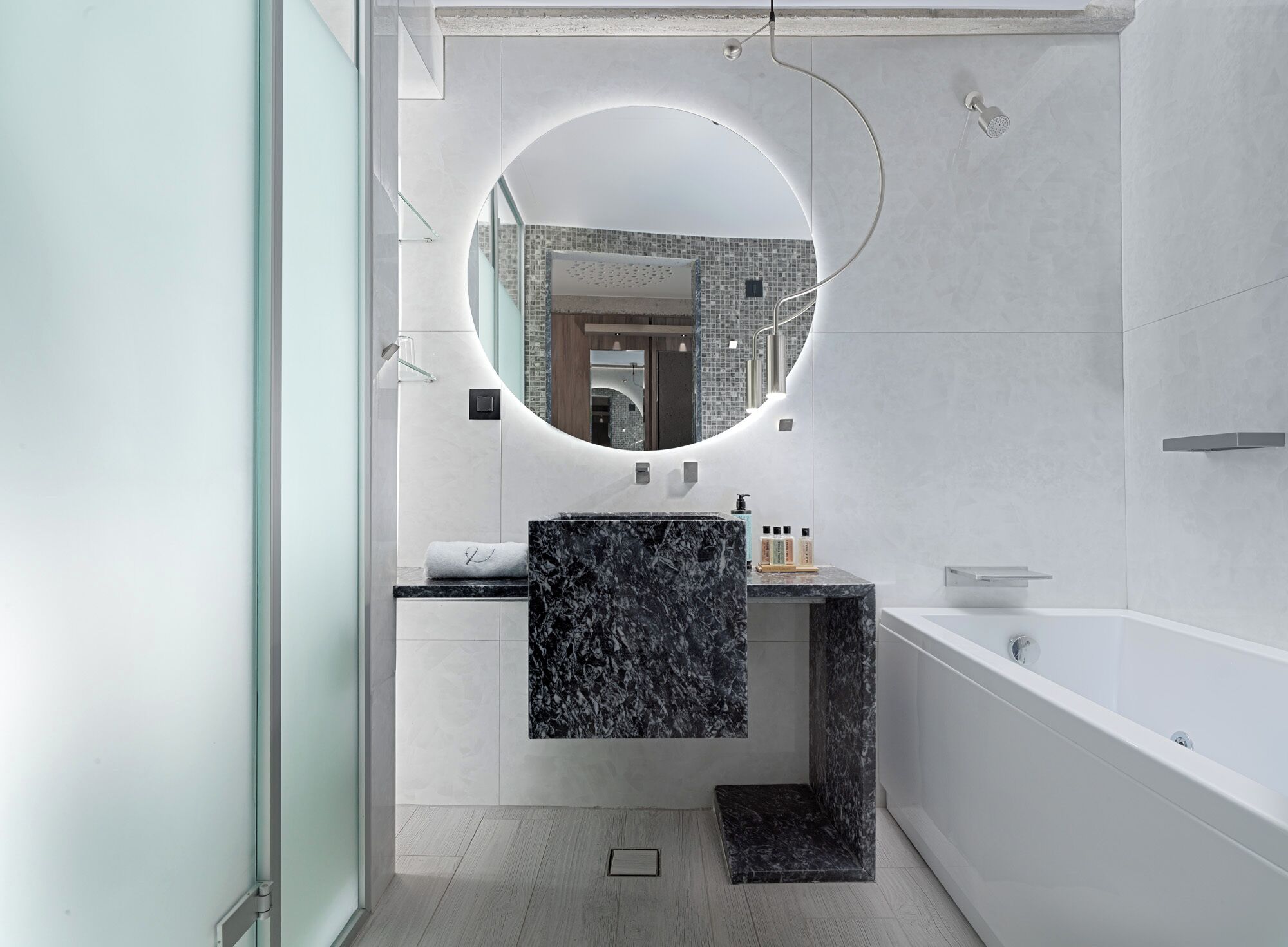 A modern bathroom with a round mirror, a marble sink, and a white bathtub.