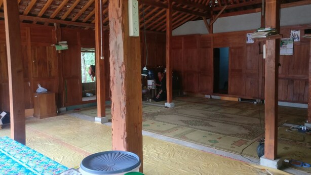 A spacious room with wooden beams and a tiled floor, featuring a table with various items and a person in the background. - Homestay An-nur (Berbah)