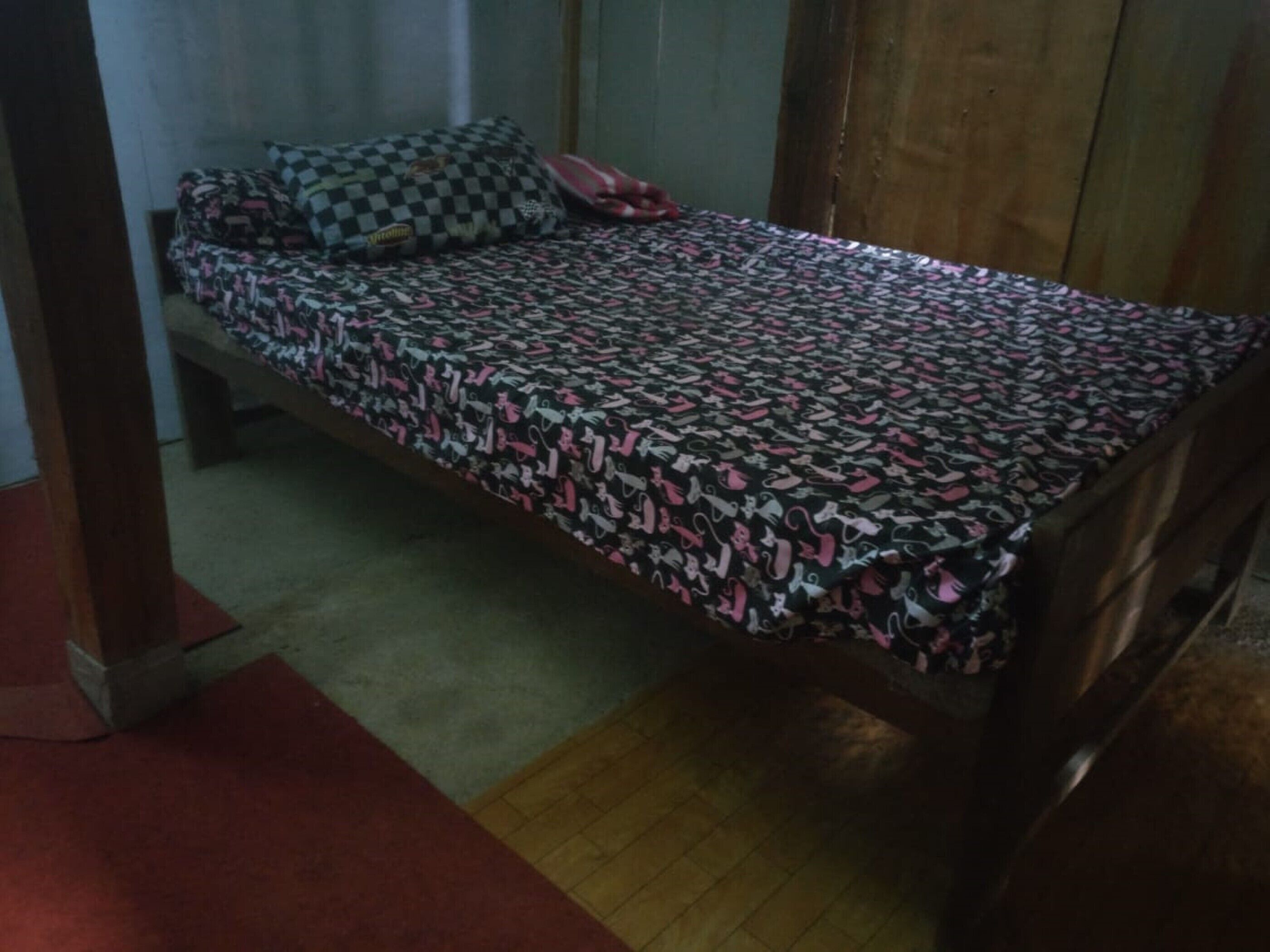 Un letto singolo con copriletto fantasia, un cuscino e una coperta rossa in una stanza con pavimento in legno e una vista parziale su una porta di legno.