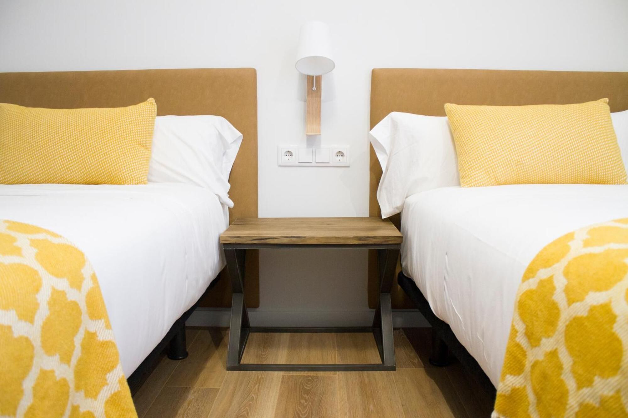 A hotel room with two beds, a nightstand, a lamp, and a power outlet on the wall.