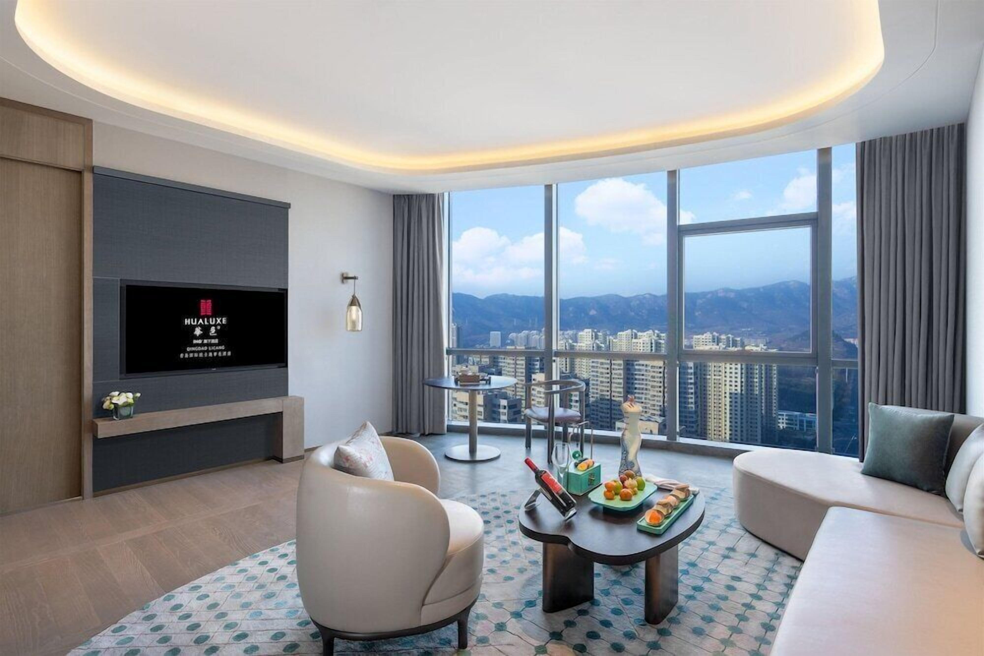 A modern hotel room with a large window offering a city view, a flat-screen TV mounted on the wall, a round coffee table with fruits and a bottle, and a sofa.