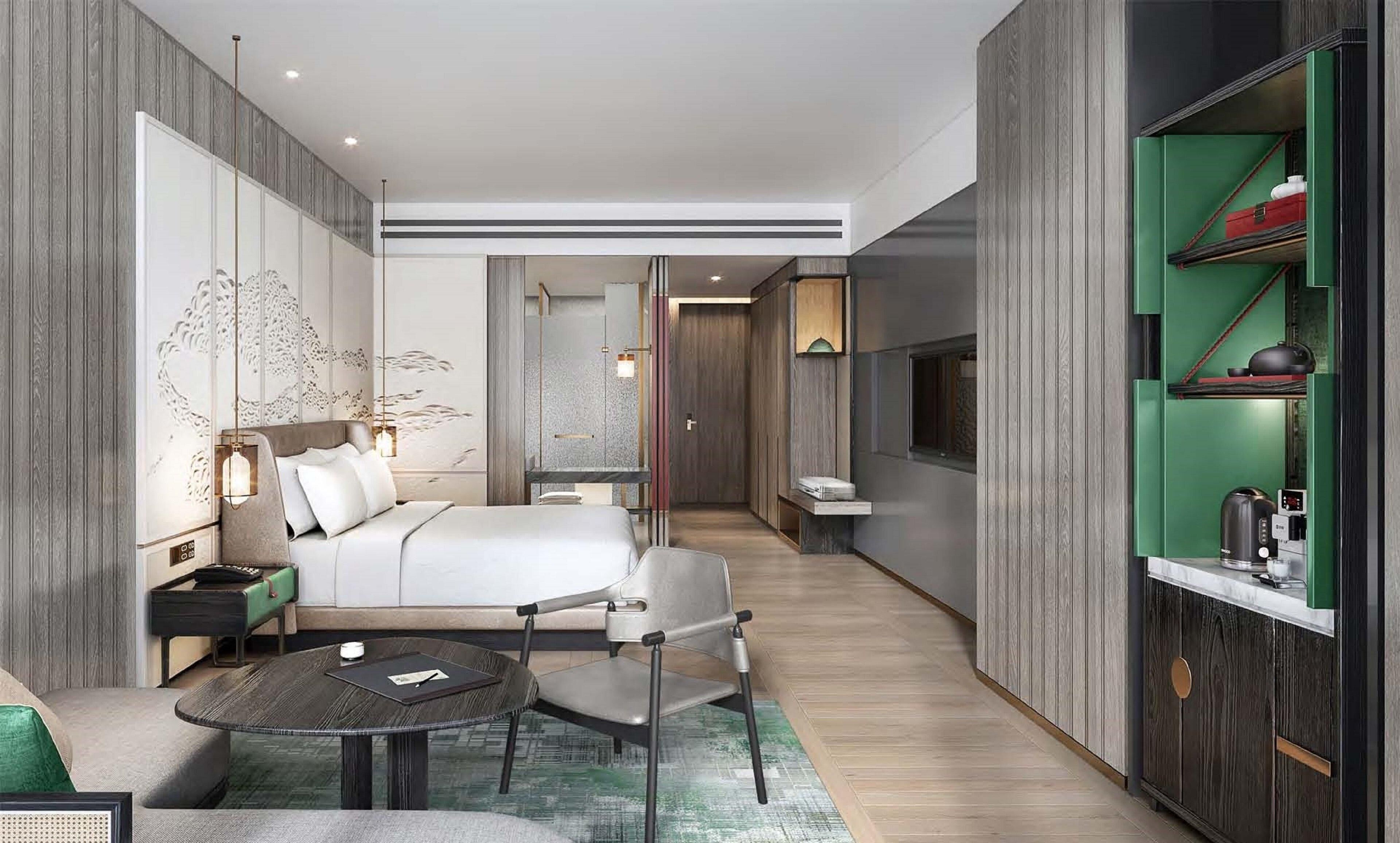 Ein modernes Hotelzimmer mit Bett, Nachttisch, Sessel und einem kleinen runden Tisch. An der Wand ist ein Fernseher angebracht und ein grünes Sideboard mit Wasserkocher darauf.