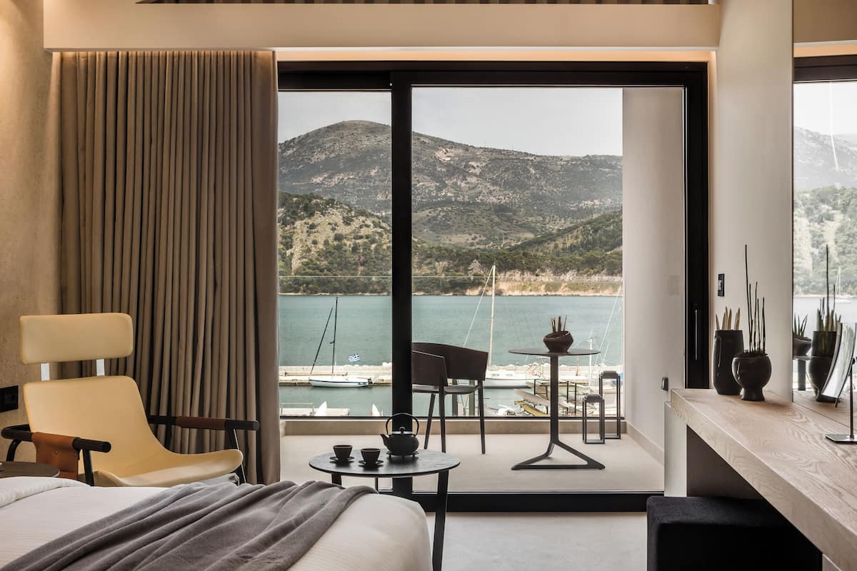 A hotel room with a bed, a chair, a small table with a teapot, and a view of a body of water and mountains.