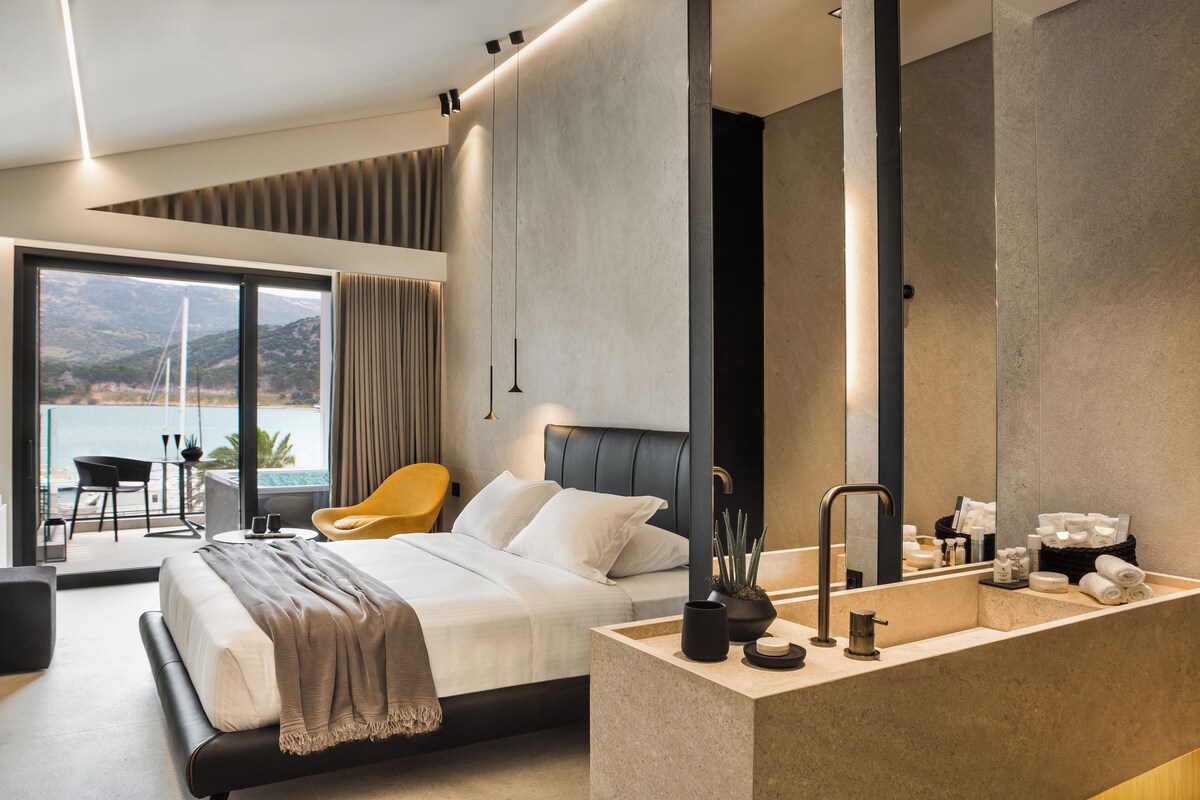A modern hotel room with a large bed, a bathroom with a large mirror, and a balcony with a view of the sea.
