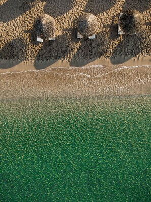 Luchtbeeld van een strand met drie rieten parasols en een ondiepe, heldere waterzone.