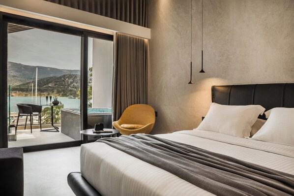 A modern hotel room with a large bed, a view of a beach and mountains, a yellow chair, and a small table. - Canale Hotel & Suites (Kefalonia)