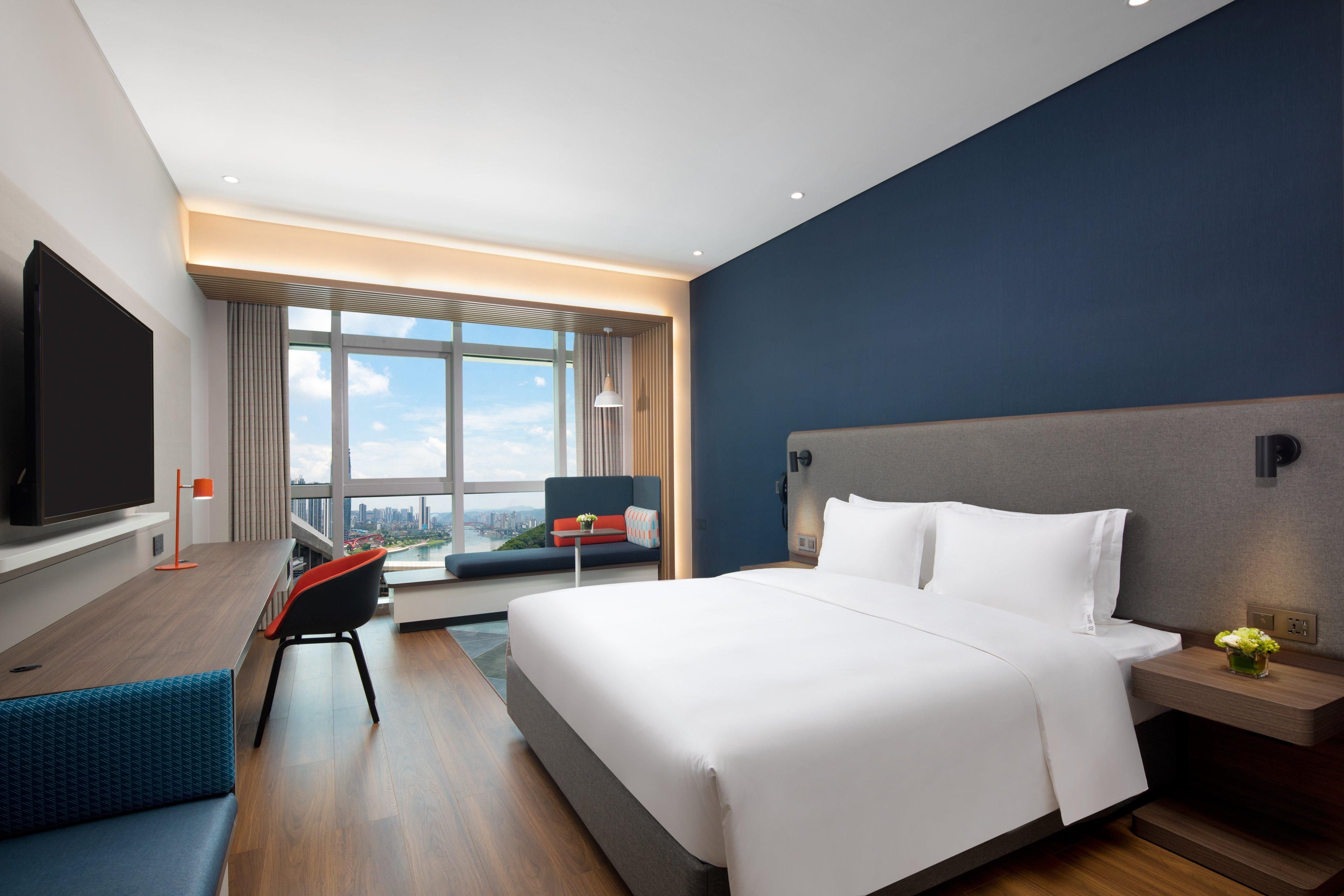Quarto de hotel moderno com uma cama grande, televisão, área de estar com vista e paisagem urbana visível pela janela.