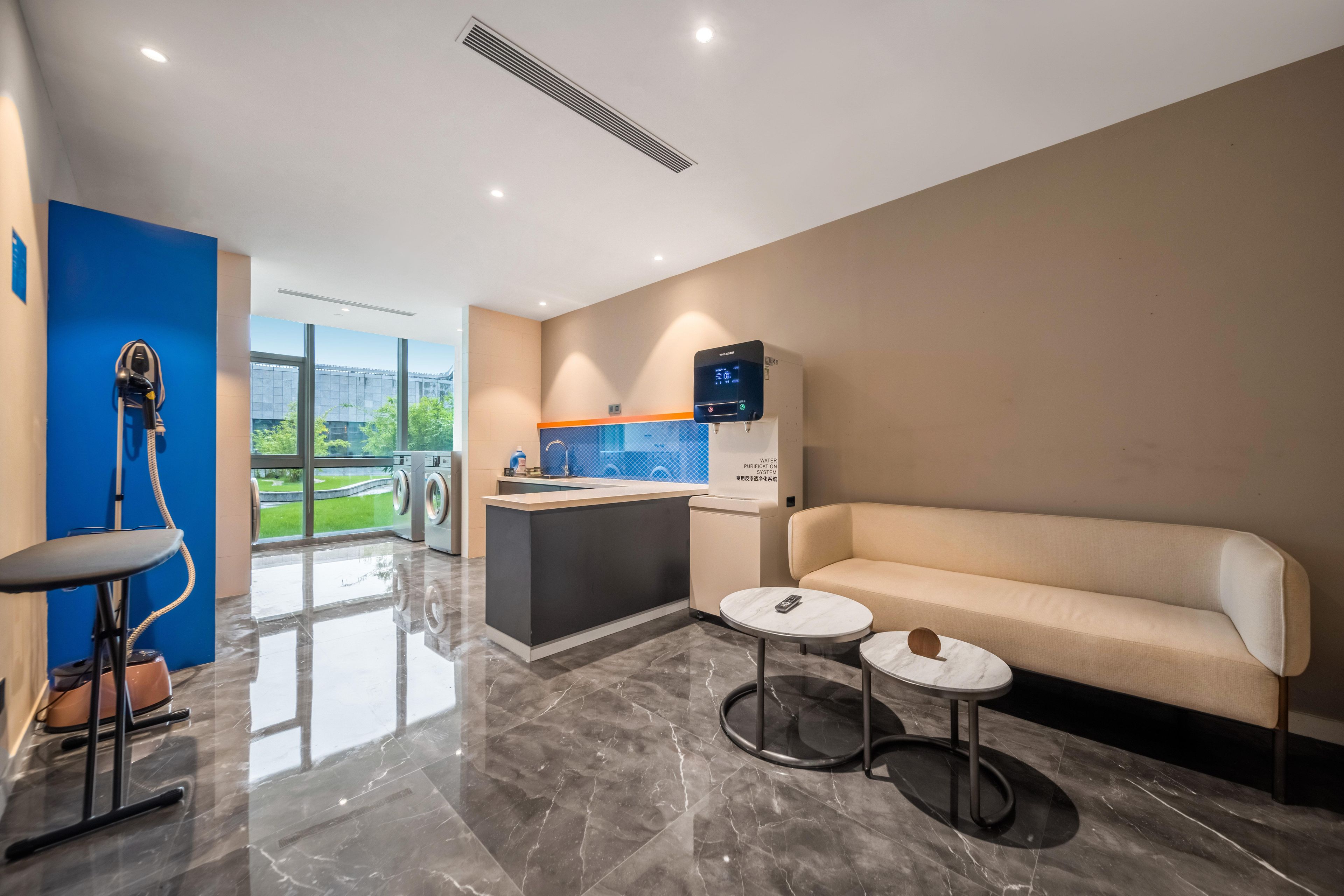 Habitación de hotel moderna con sofá, una mesita redonda, sillón de masajes, lavadora y un ventanal con vistas.