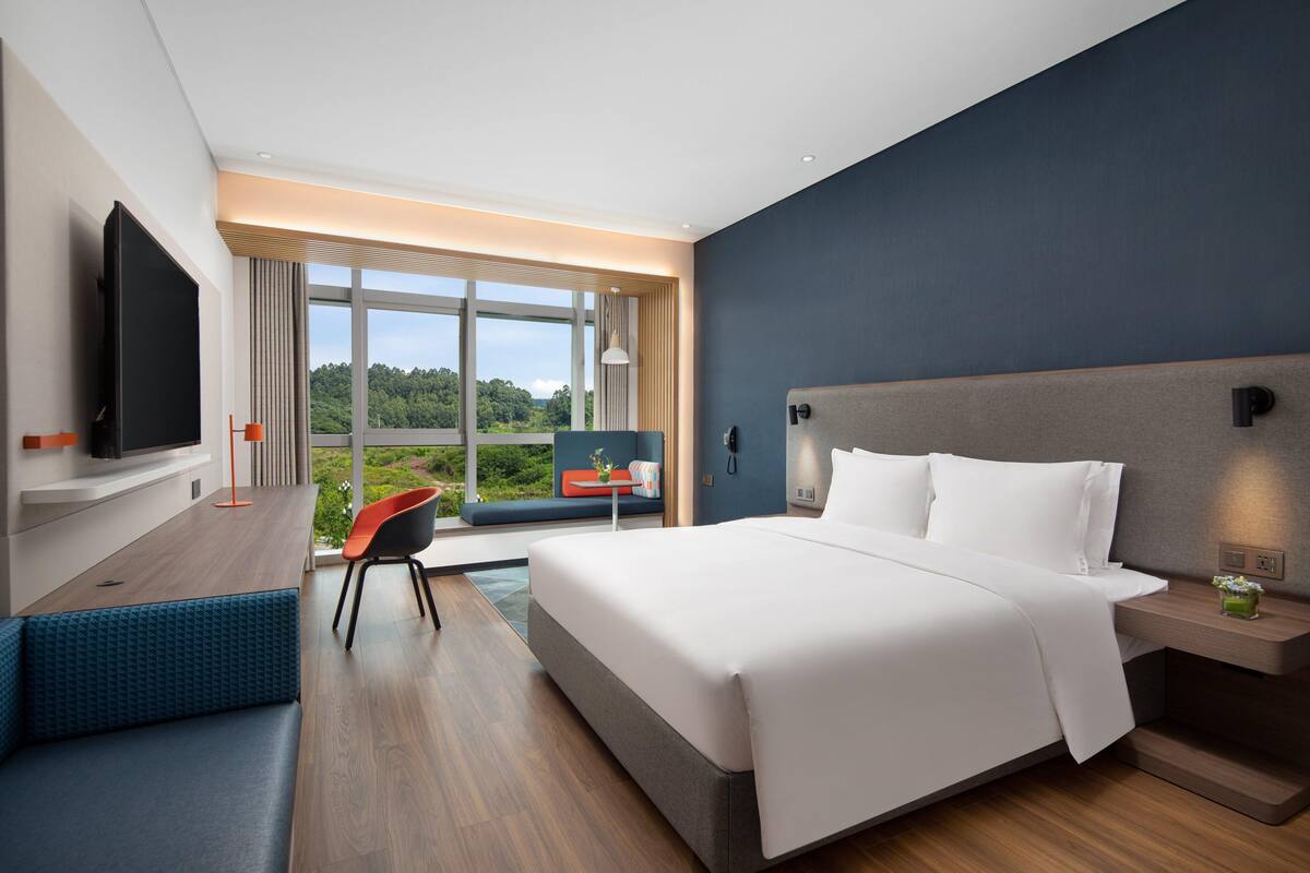 Ett modernt hotellrum med en stor säng, en TV, ett skrivbord med en stol och utsikt över grönområden utanför.
