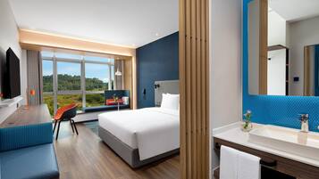 A hotel room with a bed, a desk, a chair, a large window, a bathroom with a mirror, and a sink.