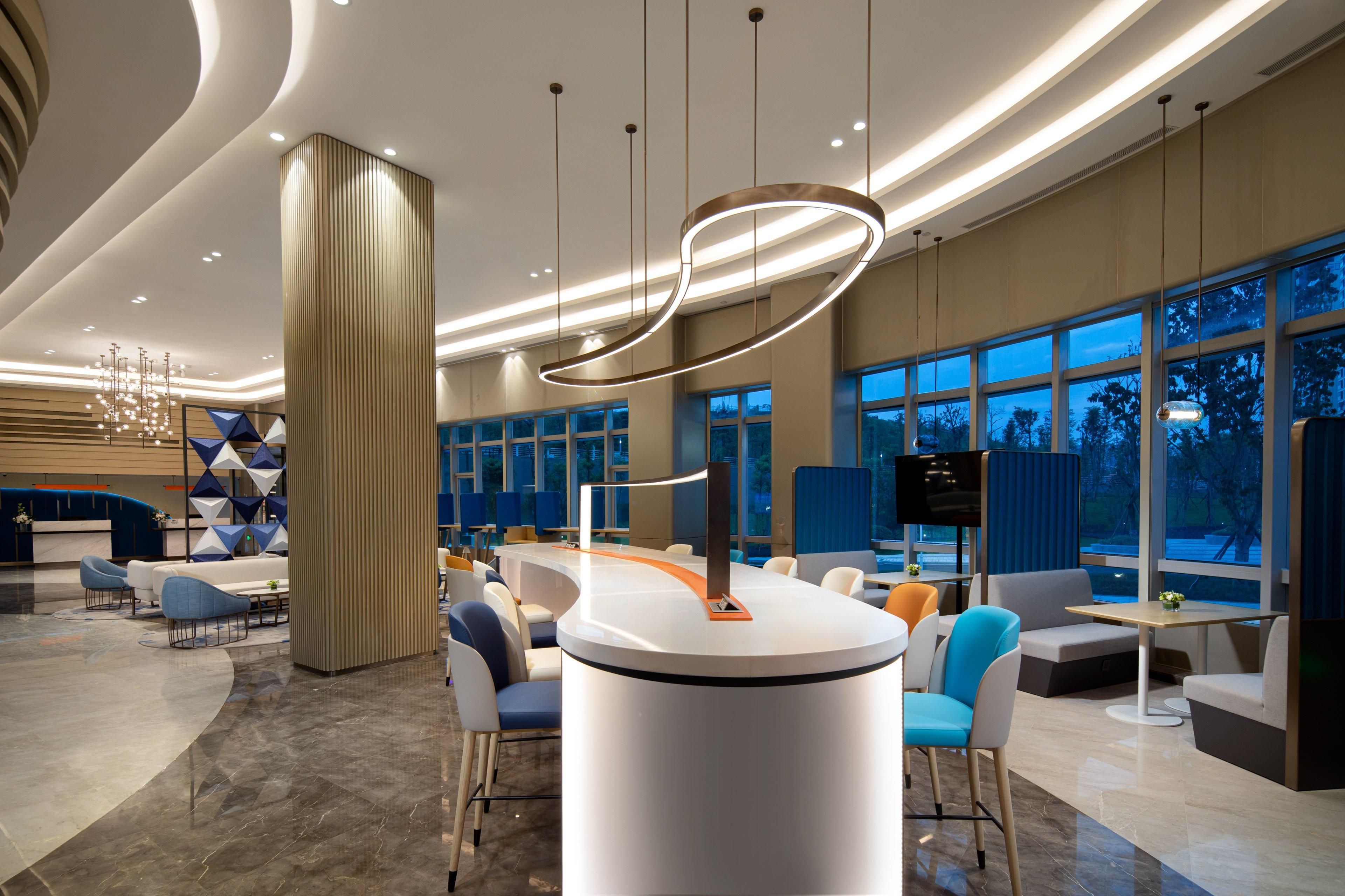 A modern hotel lobby with a central reception desk, seating areas, and large windows.