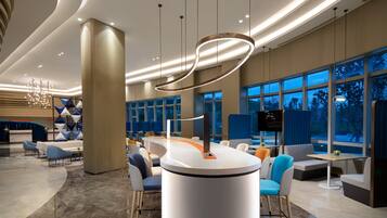 A modern hotel lobby with a central reception desk, seating areas, and large windows.