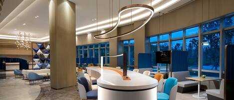 A modern hotel lobby with a central reception desk, seating areas, and large windows.