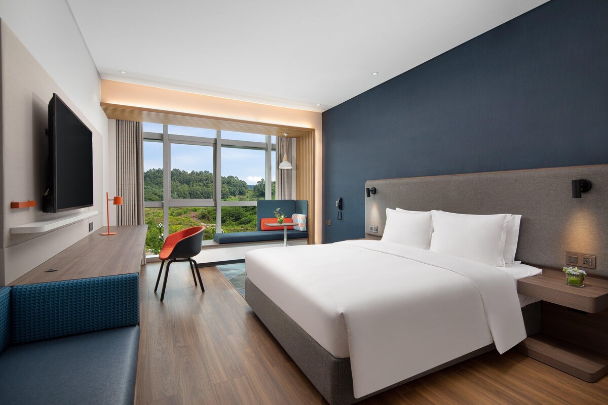 A modern hotel room with a large bed, a TV, a desk with a chair, and a view of greenery outside.