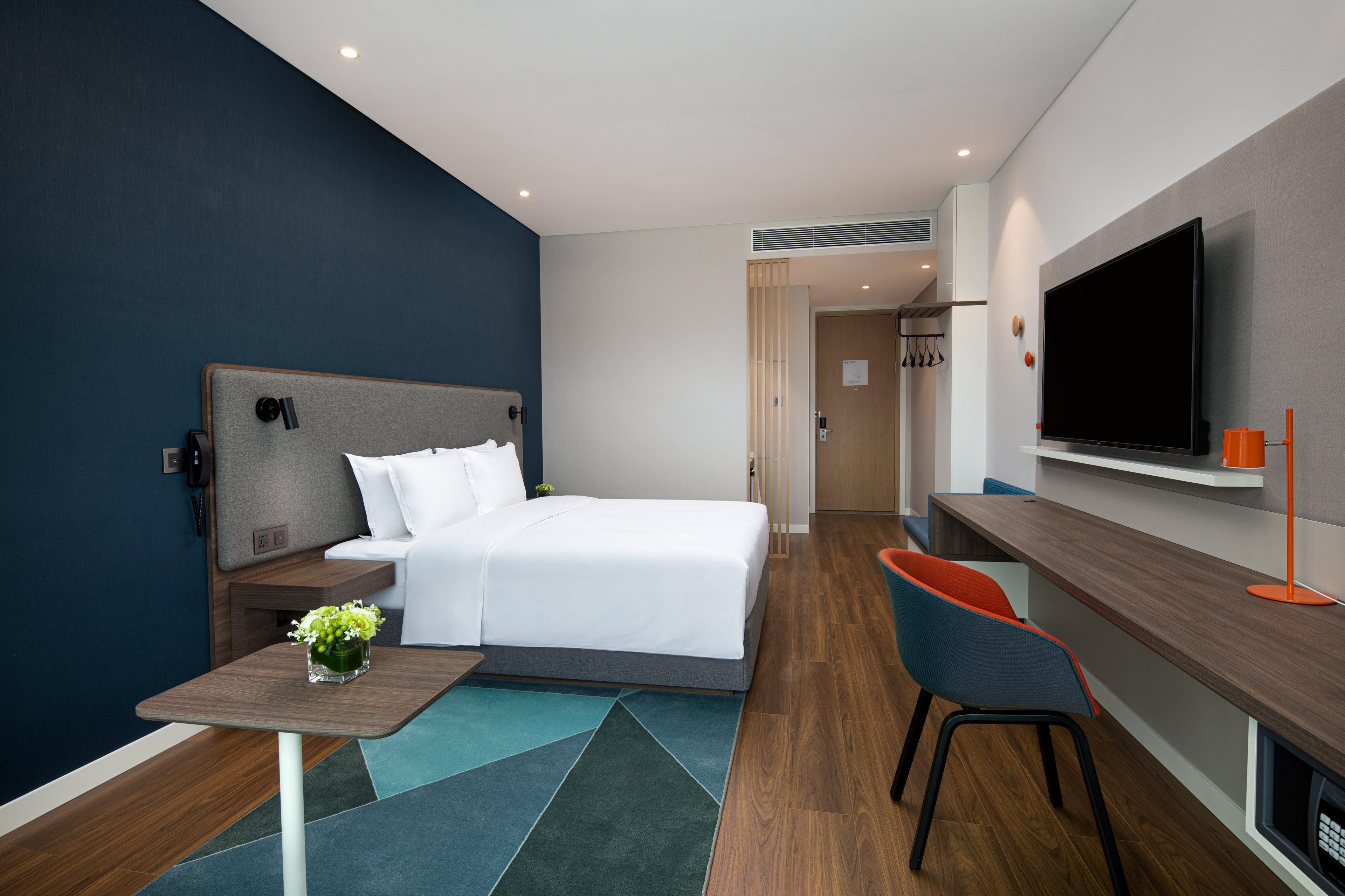 Een moderne hotelkamer met een bed, een bureau, een stoel, een televisie en een klein tafeltje met een bloevas.