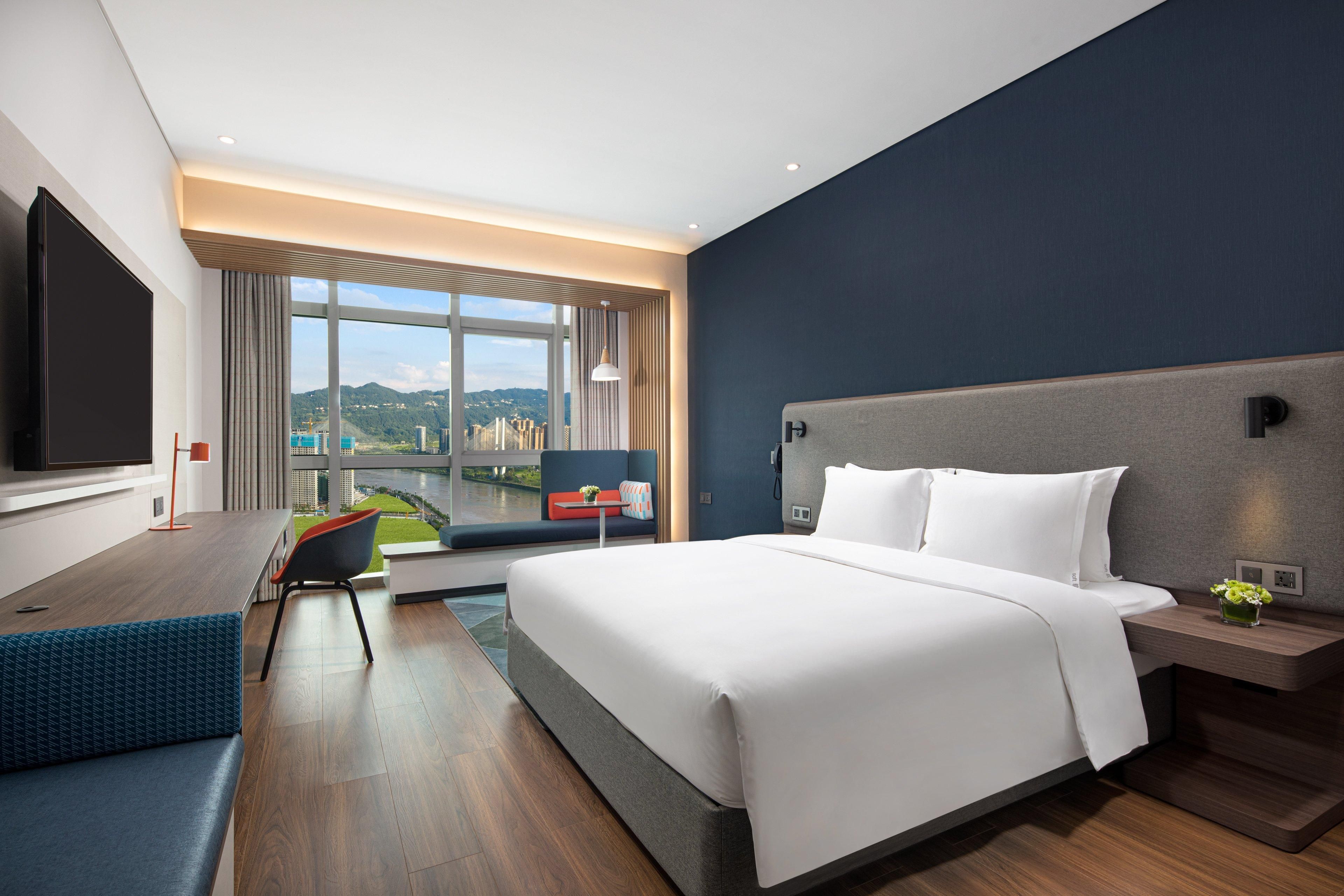 A modern hotel room with a large bed, a desk, a TV, and a view of the city.