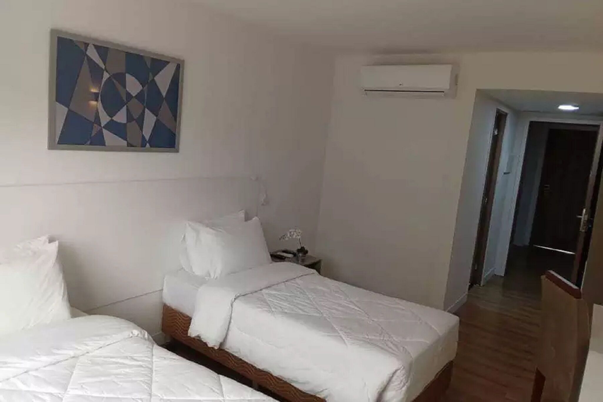 A hotel room with two beds, a wall-mounted air conditioner, and a geometric painting.