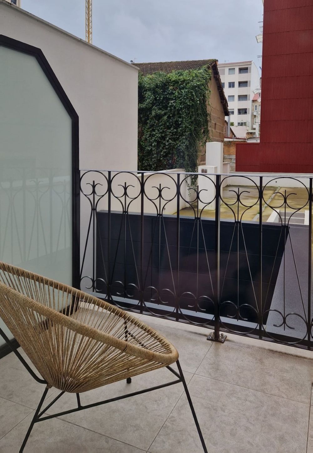 Un balcón con una silla de mimbre, una baranda de metal y vistas a un edificio con pared verde.