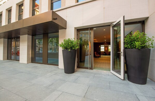 Entrada de um edifício moderno com portas de vidro, grandes vasos de plantas e um calçadão pavimentado.