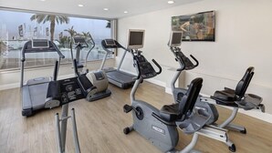 Ein Fitnessstudio mit Laufbändern, Crosstrainern und Ergometer, großen Fenstern mit Blick auf Palmen und einem Gemälde an der Wand.